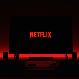 A television screen displays the Netflix logo in red against a black background. The room is dimly lit with a red glow possibly from LED backlighting behind the screen. Two speakers are positioned on either side of the TV set atop a minimalist entertainment center.