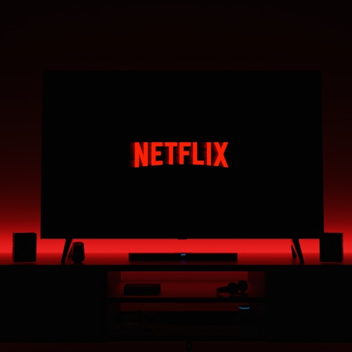 A television screen displays the Netflix logo in red against a black background. The room is dimly lit with a red glow possibly from LED backlighting behind the screen. Two speakers are positioned on either side of the TV set atop a minimalist entertainment center.