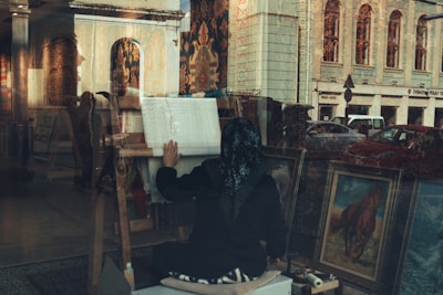 A person is weaving on a loom in a shop filled with various artworks, including framed paintings and tapestries. The scene is viewed through a window, which reflects the outside area, including cars and a large, historic building.