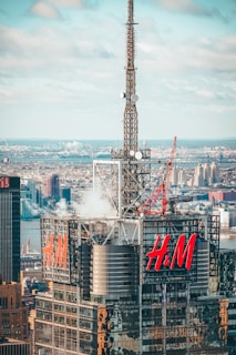 A high-rise building with a large 'H&M' logo on two sides is topped with a tall metal antenna. The cityscape in the background features numerous buildings and a river. Steam is visible coming from the rooftop, and there are cranes and various equipment present.