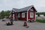 Several people are gathered around a small red wooden building with a metal roof. Some are sitting on the ground with backpacks and gear, appearing to be travelers or hikers. The building has a sign labeled 'Abisko Turiststation'. The background shows a road with a parked camper and green foliage.
