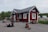 Several people are gathered around a small red wooden building with a metal roof. Some are sitting on the ground with backpacks and gear, appearing to be travelers or hikers. The building has a sign labeled 'Abisko Turiststation'. The background shows a road with a parked camper and green foliage.