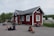 Several people are gathered around a small red wooden building with a metal roof. Some are sitting on the ground with backpacks and gear, appearing to be travelers or hikers. The building has a sign labeled 'Abisko Turiststation'. The background shows a road with a parked camper and green foliage.
