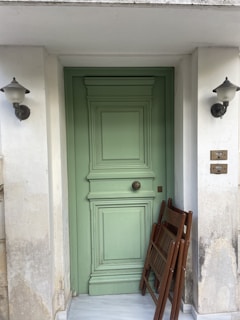 A pastel green, intricately paneled door is centered between two white walls showing signs of weathering. Two decorative wall lamps with black bases and glass tops flank the door. Leaning against the door is a wooden folding chair, adding a rustic touch. The marble flooring contrasts with the pastel tones, creating an elegant yet aged aesthetic.