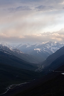 A breathtaking mountainous landscape with snow-capped peaks under a cloudy sky. A winding river meanders through the valley, with rugged terrain and lush greenery on either side.