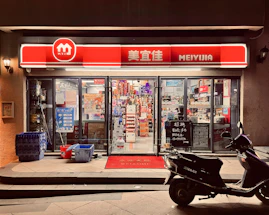 A small convenience store with a bright red and white sign that reads 'MEIYIJIA'. The store is well-lit with glass doors and windows displaying a variety of products inside, including snacks and beverages. A row of blue crates is stacked on the left side of the entrance next to some cleaning supplies. A black scooter is parked in front of the store on the right side. The pavement has a welcome mat at the entrance.
