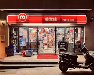 A small convenience store with a bright red and white sign that reads 'MEIYIJIA'. The store is well-lit with glass doors and windows displaying a variety of products inside, including snacks and beverages. A row of blue crates is stacked on the left side of the entrance next to some cleaning supplies. A black scooter is parked in front of the store on the right side. The pavement has a welcome mat at the entrance.