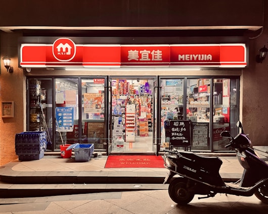 A small convenience store with a bright red and white sign that reads 'MEIYIJIA'. The store is well-lit with glass doors and windows displaying a variety of products inside, including snacks and beverages. A row of blue crates is stacked on the left side of the entrance next to some cleaning supplies. A black scooter is parked in front of the store on the right side. The pavement has a welcome mat at the entrance.