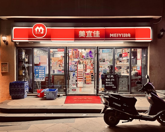A small convenience store with a bright red and white sign that reads 'MEIYIJIA'. The store is well-lit with glass doors and windows displaying a variety of products inside, including snacks and beverages. A row of blue crates is stacked on the left side of the entrance next to some cleaning supplies. A black scooter is parked in front of the store on the right side. The pavement has a welcome mat at the entrance.