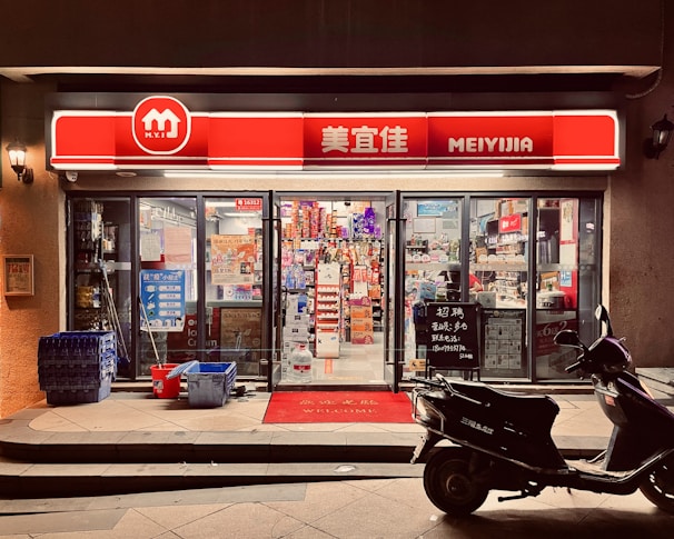 A small convenience store with a bright red and white sign that reads 'MEIYIJIA'. The store is well-lit with glass doors and windows displaying a variety of products inside, including snacks and beverages. A row of blue crates is stacked on the left side of the entrance next to some cleaning supplies. A black scooter is parked in front of the store on the right side. The pavement has a welcome mat at the entrance.