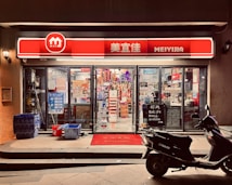 A small convenience store with a bright red and white sign that reads 'MEIYIJIA'. The store is well-lit with glass doors and windows displaying a variety of products inside, including snacks and beverages. A row of blue crates is stacked on the left side of the entrance next to some cleaning supplies. A black scooter is parked in front of the store on the right side. The pavement has a welcome mat at the entrance.