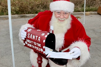 A person dressed in a Santa Claus outfit is smiling and holding a letter next to a festive mailbox labeled 'Santa Direct to the North Pole.' The mailbox is red with white snowflake decorations, and the individual appears to be in a cheerful, holiday setting.