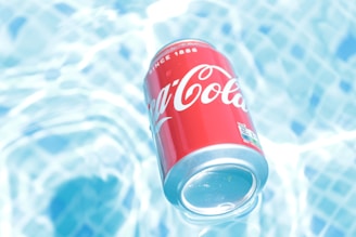 A red soda can with white lettering floats on a surface of clear, rippling water beneath it. The water is light blue and shimmers in the sunlight, creating a refreshing and cool atmosphere.