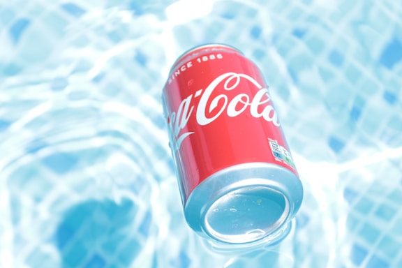 A red soda can with white lettering floats on a surface of clear, rippling water beneath it. The water is light blue and shimmers in the sunlight, creating a refreshing and cool atmosphere.