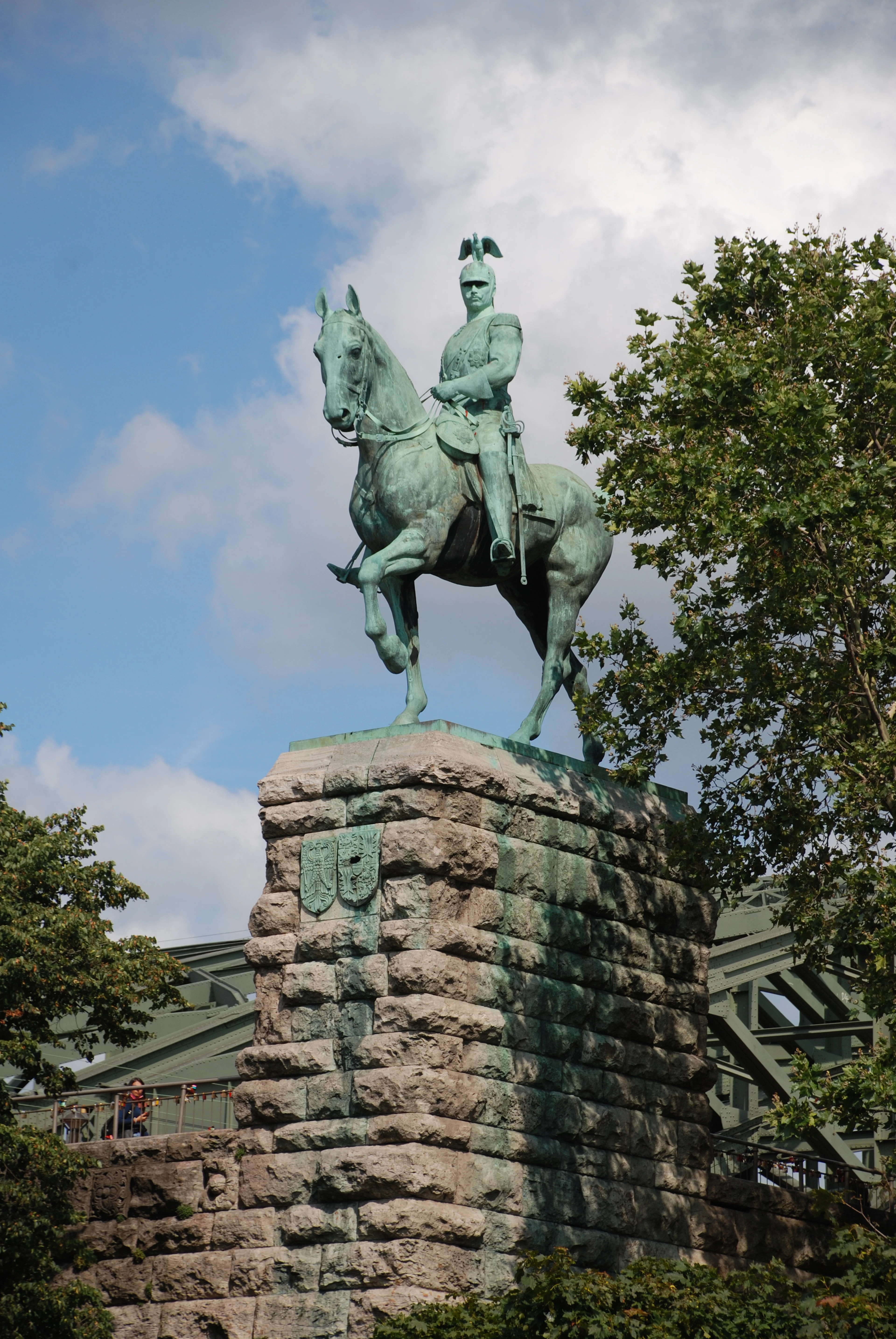 A bronze equestrian statue on a stone plinth stands in a park, framed by trees against a blue sky.