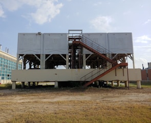 A large industrial structure with multiple white rectangular units elevated on a platform supported by pillars. A rust-colored staircase leads up to the units. The background shows a clear sky and part of a modern building with glass windows.