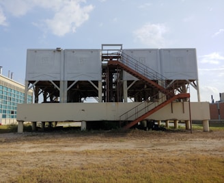 A large industrial structure with multiple white rectangular units elevated on a platform supported by pillars. A rust-colored staircase leads up to the units. The background shows a clear sky and part of a modern building with glass windows.