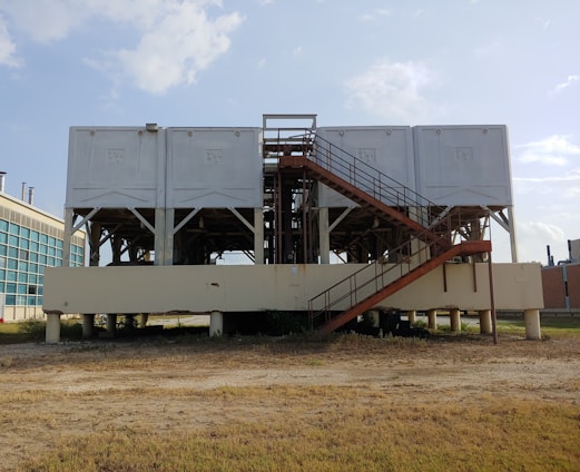 A large industrial structure with multiple white rectangular units elevated on a platform supported by pillars. A rust-colored staircase leads up to the units. The background shows a clear sky and part of a modern building with glass windows.