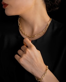 A person is wearing a gold chain necklace and matching bracelet. The individual is dressed in black, and the focus is on the luxurious jewelry. The hand is elegantly posed near the neck, highlighting both the bracelet and necklace.