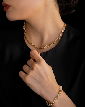 A person is wearing a gold chain necklace and matching bracelet. The individual is dressed in black, and the focus is on the luxurious jewelry. The hand is elegantly posed near the neck, highlighting both the bracelet and necklace.