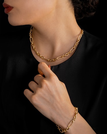 A person is wearing a gold chain necklace and matching bracelet. The individual is dressed in black, and the focus is on the luxurious jewelry. The hand is elegantly posed near the neck, highlighting both the bracelet and necklace.