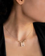 A person wearing a necklace featuring a gold padlock pendant. The padlock is inscribed with 'Tiffany & Co.' and is attached to a delicate chain that is visible against the person's neck. The background is dark, contrasting with the warm tones of the skin and the gold jewelry.
