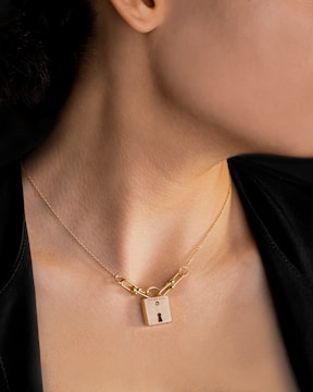 A person wearing a necklace featuring a gold padlock pendant. The padlock is inscribed with 'Tiffany & Co.' and is attached to a delicate chain that is visible against the person's neck. The background is dark, contrasting with the warm tones of the skin and the gold jewelry.