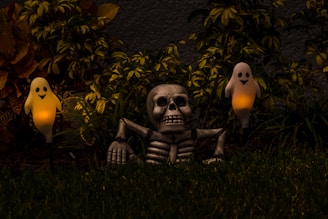 A Halloween-themed setup featuring a skeleton partially buried in the grass with two glowing ghost figures on either side. The scene is set against a backdrop of dark foliage, with the glowing ghosts providing an eerie ambiance to the night setting.