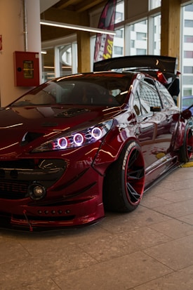 A modified, sleek sports car with a glossy deep red finish is parked indoors. The vehicle features eye-catching LED headlights and wide, custom rims. Large windows and a raised spoiler add to its stylish appearance. The setting appears to be a showroom with bright lights and large windows in the background.