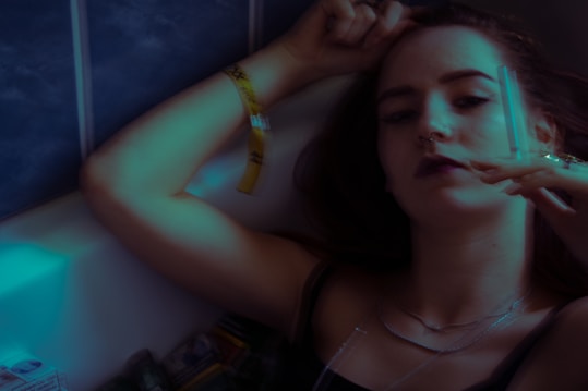 A person with a nose ring is reclining, holding what appears to be a syringe or similar object. The dimly lit setting gives a moody atmosphere, and the person is wearing a yellow wristband and multiple necklaces.