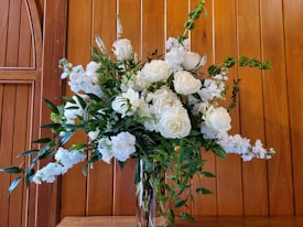 A floral arrangement features white roses and other flowers with lush green foliage, placed in a clear glass vase against a background of wooden paneling with a warm tone.