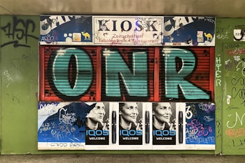A storefront with vibrant graffiti covers the front of a kiosk. Large, bold letters 'ONR' are painted in teal and black across a shuttered entrance. Surrounding the letters are various posters and stickers, including adjacent black and white advertisements of a smiling person with the text 'IQOS WELCOME'. The walls are lined with various tags and graffiti in multiple colors.