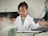 A young child wearing a white lab coat and safety goggles is standing in front of a table, appearing to conduct a science experiment. A metal container emits a misty vapor, creating a sense of activity and curiosity. The background features a neutral wall with blurred elements, such as orange stools and a tripod stand.