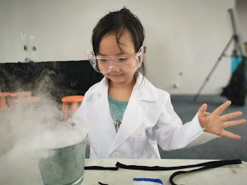 A young child wearing a white lab coat and safety goggles is standing in front of a table, appearing to conduct a science experiment. A metal container emits a misty vapor, creating a sense of activity and curiosity. The background features a neutral wall with blurred elements, such as orange stools and a tripod stand.
