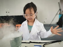 A young child wearing a white lab coat and safety goggles is standing in front of a table, appearing to conduct a science experiment. A metal container emits a misty vapor, creating a sense of activity and curiosity. The background features a neutral wall with blurred elements, such as orange stools and a tripod stand.