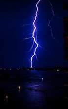 A vivid lightning bolt strikes from the sky, illuminating the dark, overcast night. The water below reflects the bright flash, and small points of light are visible in the distance, suggesting city lights or reflections on the water's surface. A silhouette of a building is partially visible on the right edge.