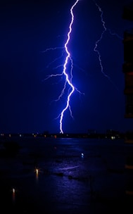 A vivid lightning bolt strikes from the sky, illuminating the dark, overcast night. The water below reflects the bright flash, and small points of light are visible in the distance, suggesting city lights or reflections on the water's surface. A silhouette of a building is partially visible on the right edge.