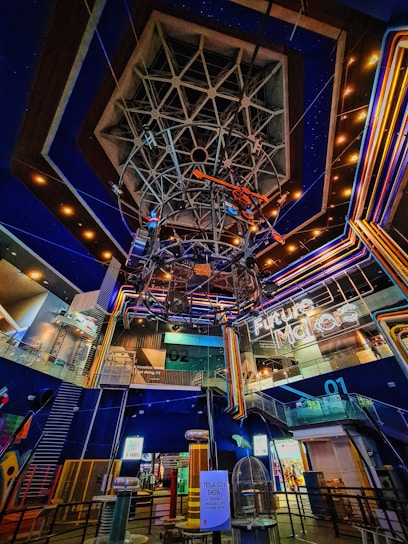 A futuristic indoor setting with a large geometric structure hanging from the ceiling and neon lights. The area showcases vibrant, multicolored lighting with signs indicating a 'Future Makers' exhibit and a Tesla coil show. The environment suggests a science or technology museum.
