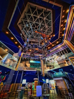 A futuristic indoor setting with a large geometric structure hanging from the ceiling and neon lights. The area showcases vibrant, multicolored lighting with signs indicating a 'Future Makers' exhibit and a Tesla coil show. The environment suggests a science or technology museum.