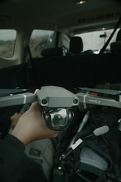 A hand is holding a drone, specifically a DJI Mavic 2, inside a vehicle. The drone's camera is visible, and the interior of the vehicle, including seats and seatbelts, can be seen in the background.
