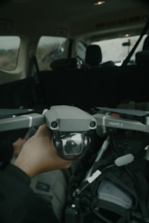 A hand is holding a drone, specifically a DJI Mavic 2, inside a vehicle. The drone's camera is visible, and the interior of the vehicle, including seats and seatbelts, can be seen in the background.