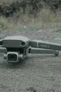 A drone labeled 'Mavic 2' with a Hasselblad camera rests on a textured asphalt surface. The background features blurred grass and rocks.