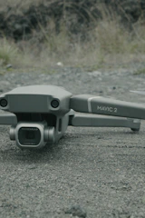 A drone labeled 'Mavic 2' with a Hasselblad camera rests on a textured asphalt surface. The background features blurred grass and rocks.