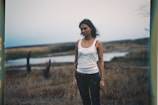A person with tattoos stands in a natural, open field landscape with tall grasses and a body of water in the background. The sky appears overcast, contributing to a moody atmosphere.