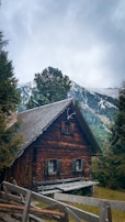 A rustic wooden cabin nestled among trees in a mountainous landscape. Snow-capped peaks rise in the background under a cloudy sky, while a simple wooden fence leads up to the cabin, adding to the cozy, secluded atmosphere.