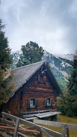A rustic wooden cabin nestled among trees in a mountainous landscape. Snow-capped peaks rise in the background under a cloudy sky, while a simple wooden fence leads up to the cabin, adding to the cozy, secluded atmosphere.