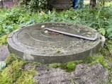 A round concrete manhole cover with circular grooves is placed on a moss-covered surface. It is surrounded by overgrown vegetation, and a metal crowbar rests on top of the cover.
