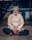 An elderly man with white hair and a bushy beard sits cross-legged on the ground, wearing a light beige tunic and dark pants. His eyes are closed, and he appears to be meditating or in deep concentration. A red tilak and white markings are on his forehead, signifying a religious or spiritual practice. The background is slightly blurred, with a building and some greenery visible.
