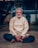 An elderly man with white hair and a bushy beard sits cross-legged on the ground, wearing a light beige tunic and dark pants. His eyes are closed, and he appears to be meditating or in deep concentration. A red tilak and white markings are on his forehead, signifying a religious or spiritual practice. The background is slightly blurred, with a building and some greenery visible.