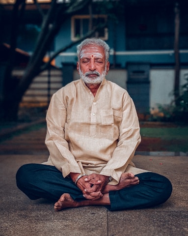 An elderly man with white hair and a bushy beard sits cross-legged on the ground, wearing a light beige tunic and dark pants. His eyes are closed, and he appears to be meditating or in deep concentration. A red tilak and white markings are on his forehead, signifying a religious or spiritual practice. The background is slightly blurred, with a building and some greenery visible.